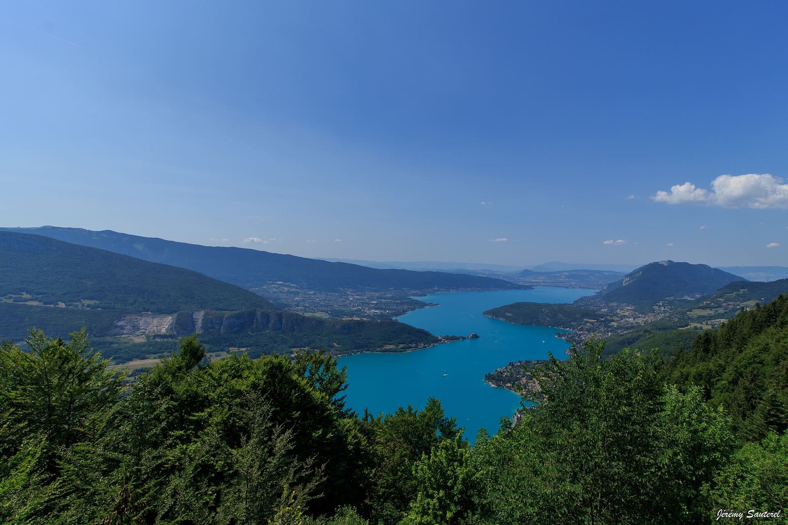 Le lac d’Annecy turquoise vu d’en haut, entre des pentes boisées
